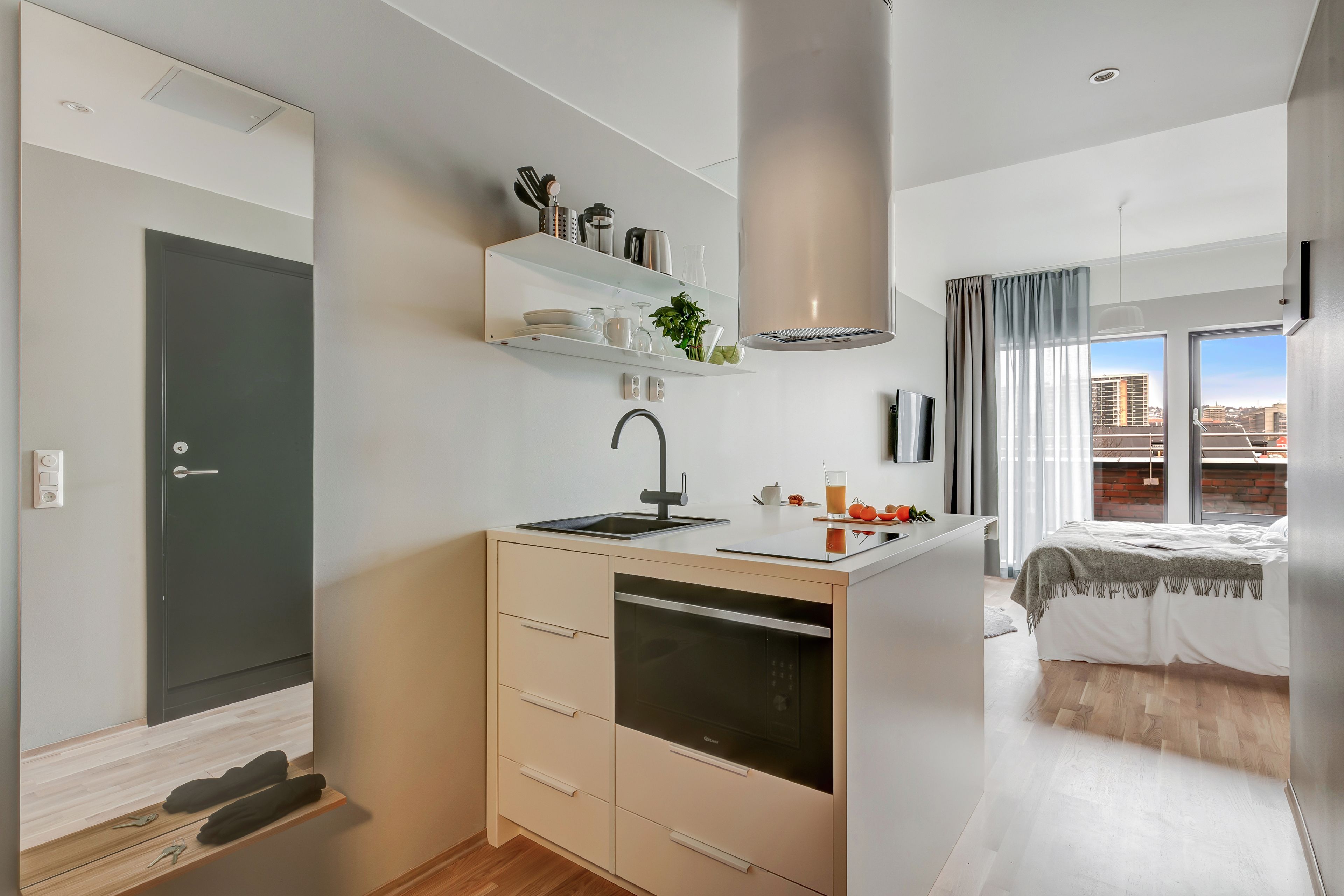 A modern kitchen with a dishwasher, a sink, and a countertop with kitchen utensils. There is a bed with a white comforter and a view of the city through large windows.