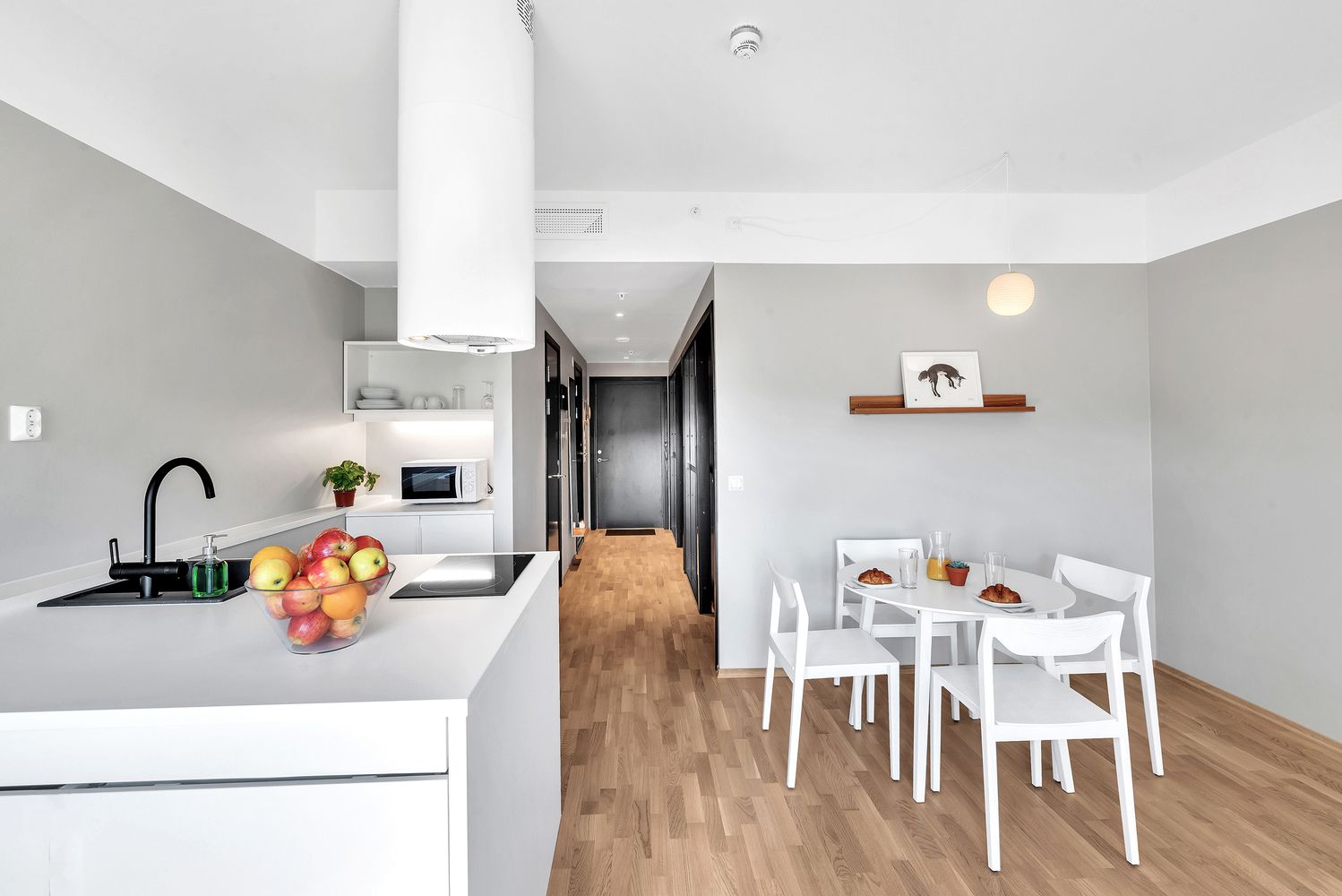 Une cuisine moderne avec un plan de travail blanc, un évier et un four à micro-ondes. On y trouve un coin repas avec des chaises et une table blanches, ainsi qu’un couloir menant à une autre pièce.