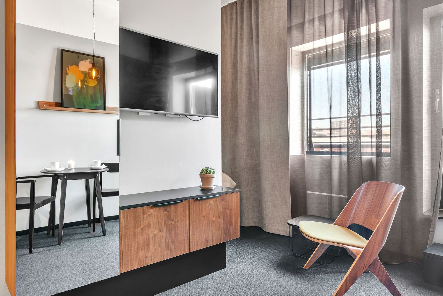 A modern hotel room with a flat-screen TV mounted on the wall, a small dining table with chairs, a wooden cabinet, a potted plant, and a large window with curtains.