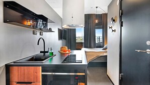 Cocina compacta con lavamanos, estufa y una pequeña encimera. Se observa una cama con vista a la ciudad de fondo.