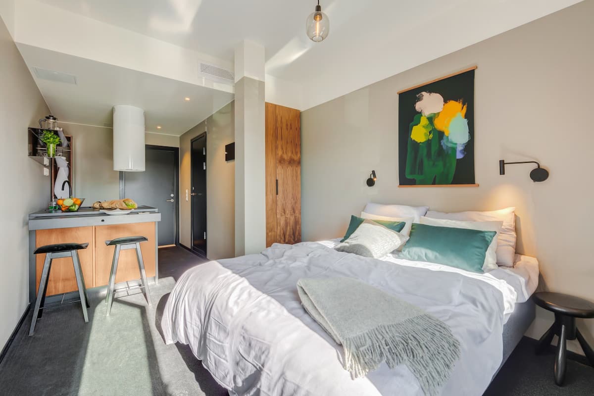 A modern bedroom with a large bed, a small kitchenette, and a painting on the wall.