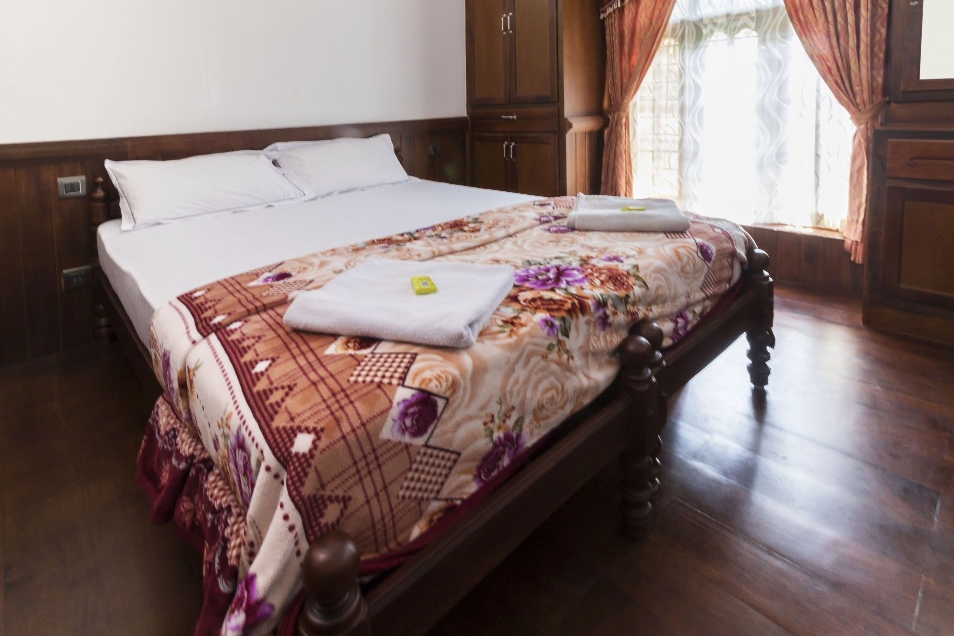 A neatly made bed with a floral bedspread, wooden headboard, and a small bedside table with a towel.