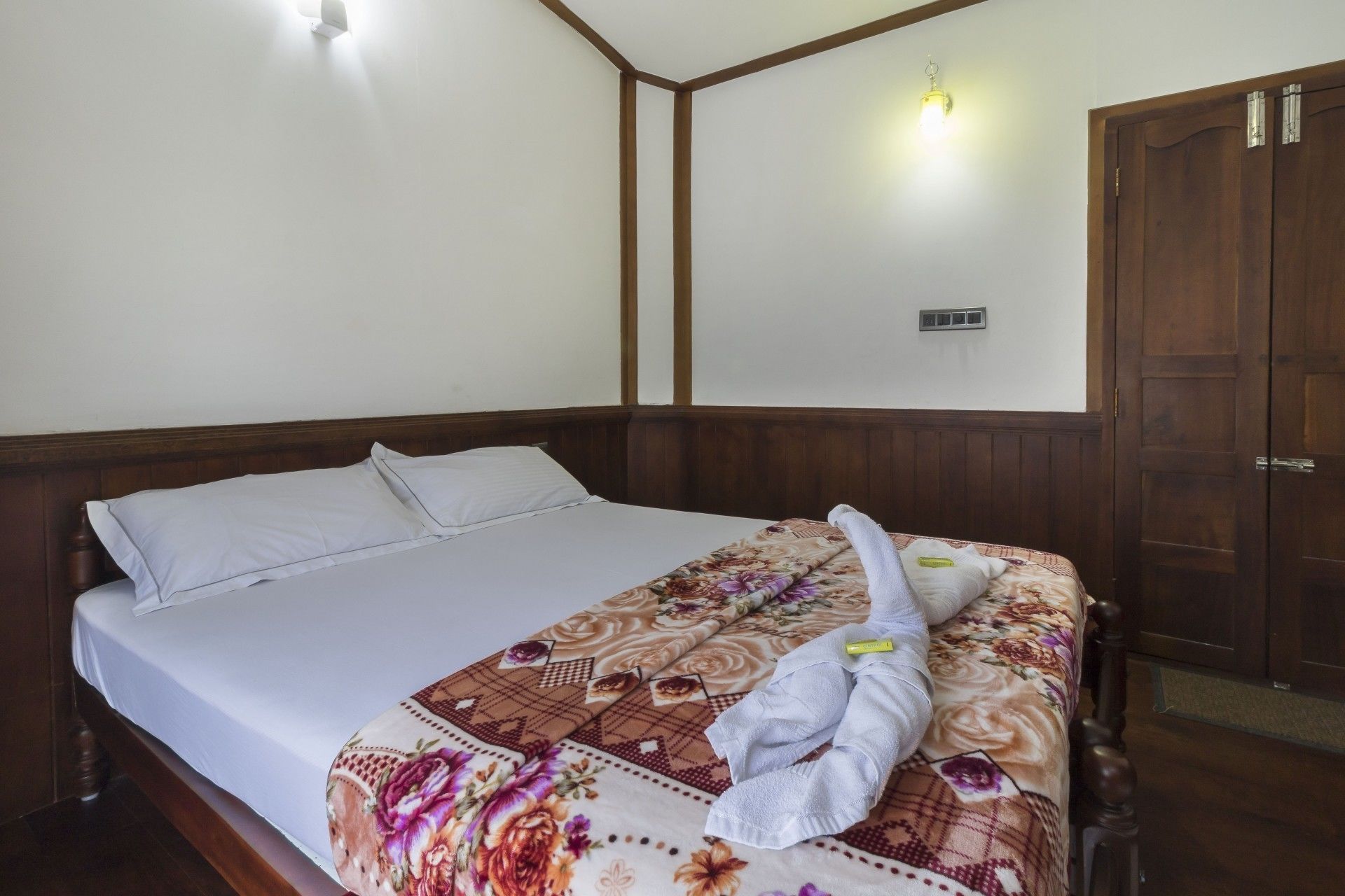 A neatly made bed with a floral bedspread and a white towel, wooden headboard, and a wooden wardrobe with a door.