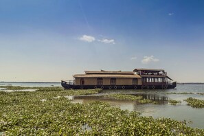 A houseboat with a flat roof and multiple windows, floating on a body of water surrounded by lily pads. - GuestHouser 3 BHK Houseboat 1b08 (Kottayam)