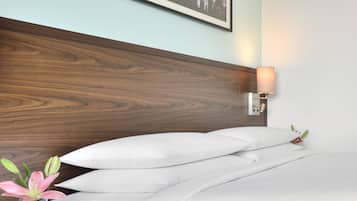 A neatly made bed with white linens, a wooden headboard, a framed picture above the bed, a bedside table with a vase of flowers, and a wall-mounted lamp.