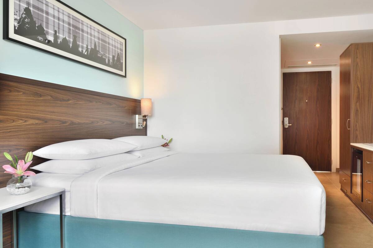 Un lit bien fait, avec du linge de lit blanc, une tête de lit en bois, une lampe fixée au mur et un tableau encadré au-dessus.
