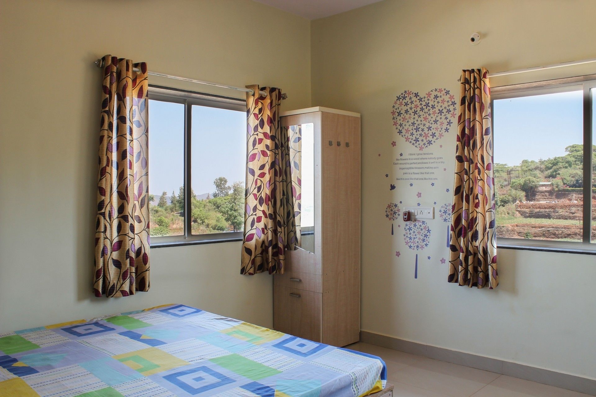 Une pièce comprenant un lit, deux fenêtres avec des rideaux, une armoire et un mur orné d’un motif floral.