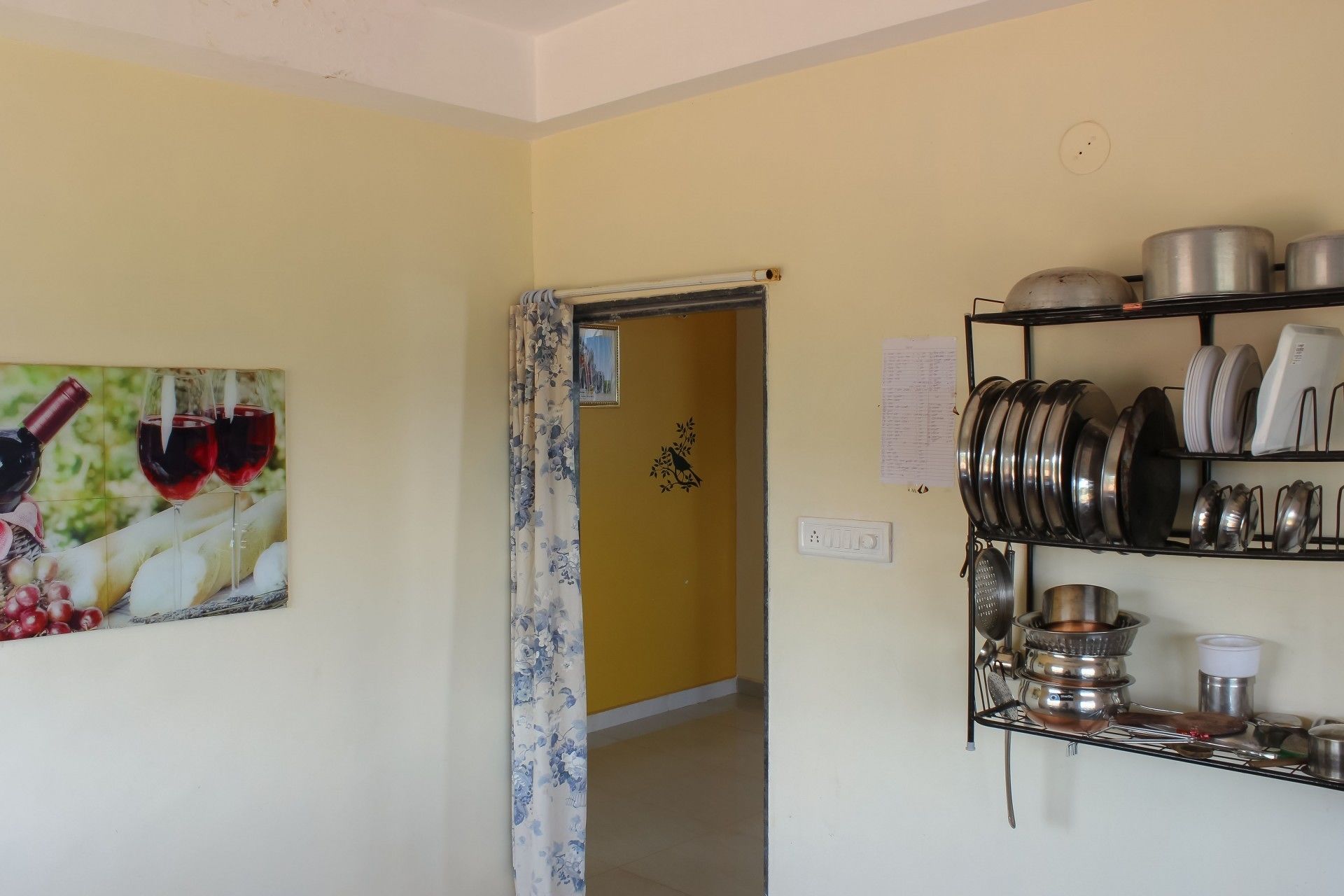 A kitchen with a metal rack holding pots and pans, a door leading to another room, and a wall with a framed picture of wine bottles.