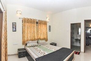 Quarto com uma cama, duas mesas de cabeceira, um criado-mudo, uma janela com cortinas e um banheiro visível por uma porta aberta.