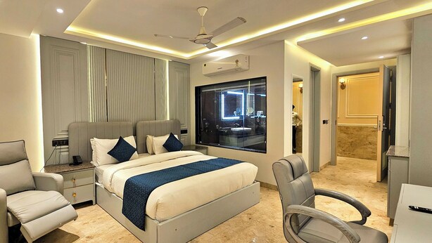 Una habitación de hotel moderna con una cama grande, dos sillones, una mesita de noche, un tocador con espejo y una ventana con vistas a la ciudad.