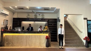 Un mostrador de recepción con un cartel que dice "RECEPTION", un reloj y una estatua de Buda. Hay tres personas detrás del mostrador y una escalera a la derecha.