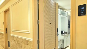 Un pasillo de hotel con suelo de mármol, una puerta con panel texturizado y una cocina visible a través de otra puerta abierta.