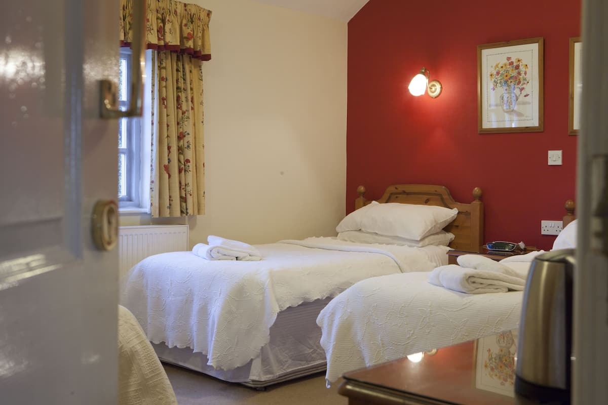 A bedroom with two beds, a window with floral curtains, a wall-mounted light, and a framed picture.