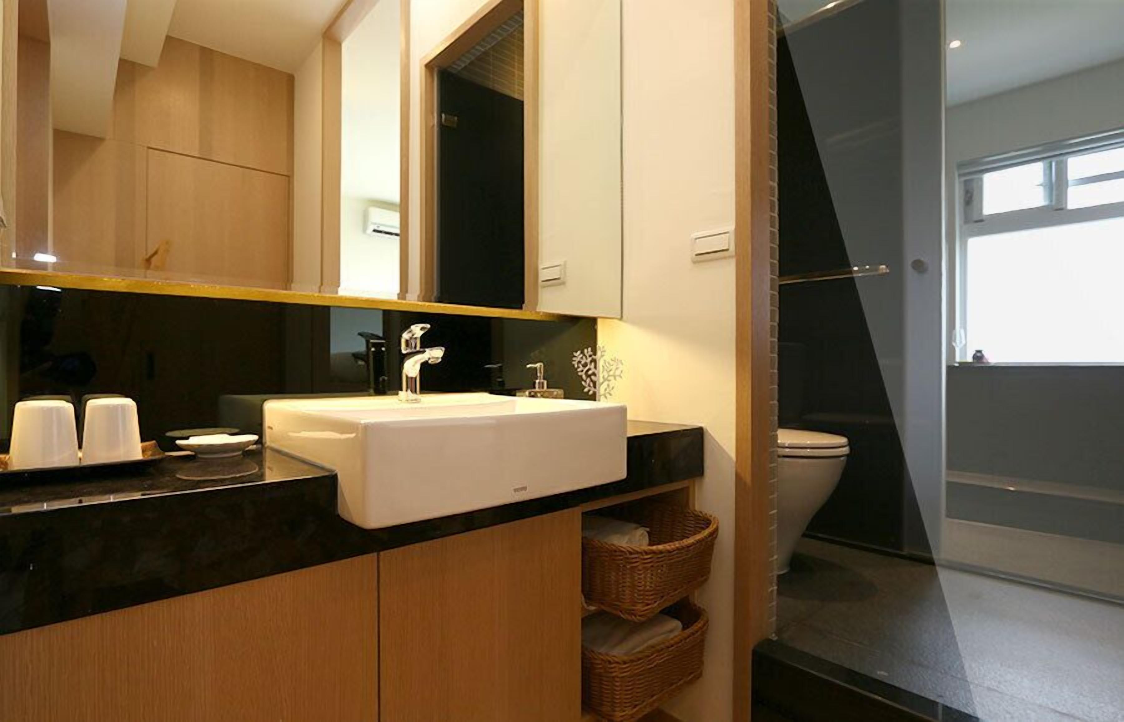 A modern bathroom with a black countertop, white sink, and a large mirror. There are wicker baskets under the sink and a toilet visible in the background.