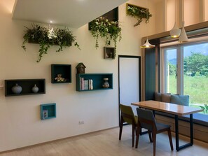 A modern dining area with a wooden table and chairs, surrounded by green shelves with plants and books.