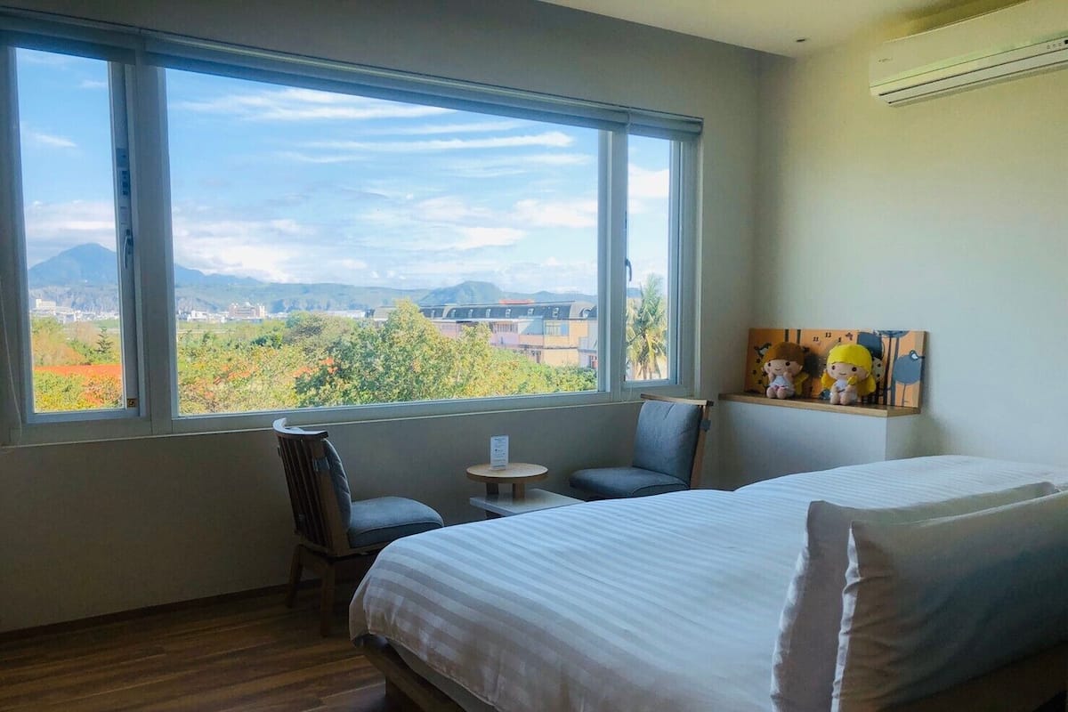 A hotel room with a large window offering a view of a cityscape and mountains. There are two beds, a small table, and a chair.