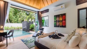 A modern bedroom with a large bed, a desk, and a chair. There is a view of a swimming pool and greenery outside. - Inspire Villas Phuket (Rawai)