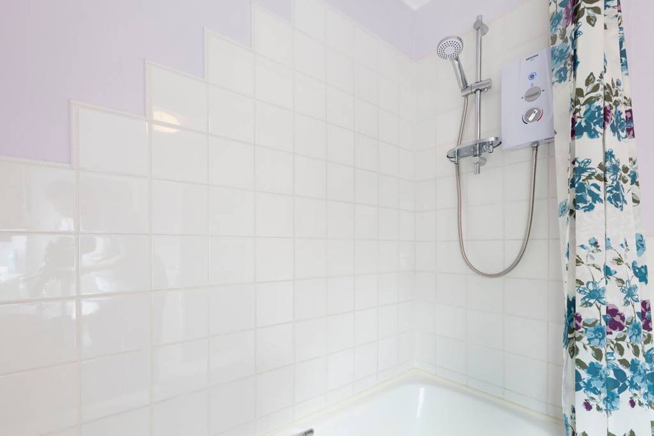 A bathroom with white tiled walls, a shower with a handheld showerhead, and a floral shower curtain.