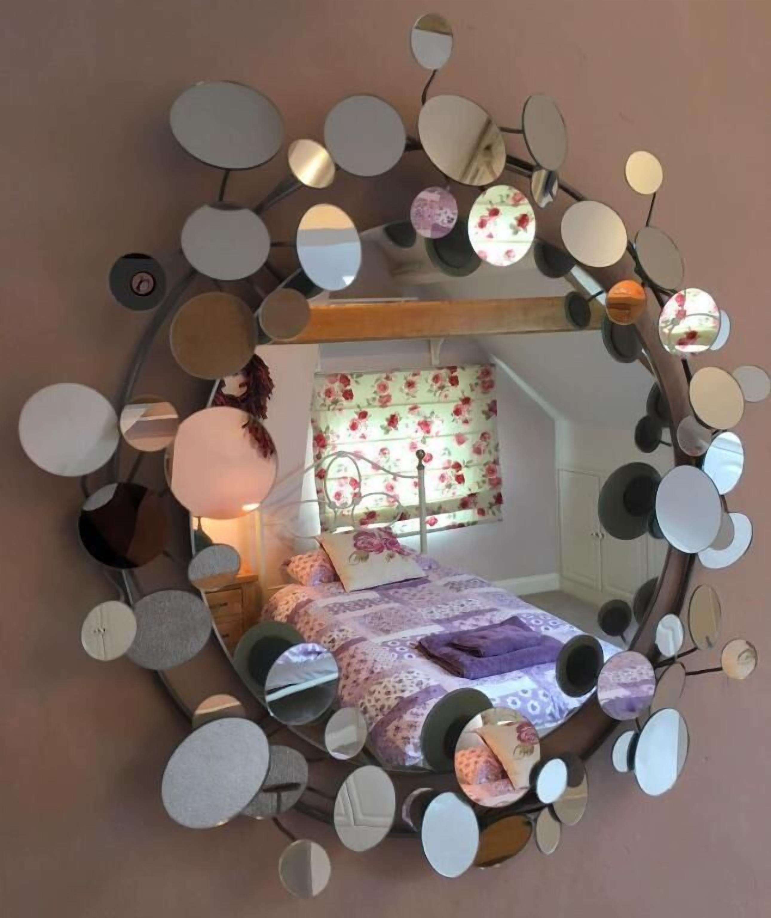 Een ronde spiegel met meerdere kleinere spiegels eraan bevestigd, die een slaapkamer weerspiegelt met een bed, kussens en een raam met bloemen gordijnen.