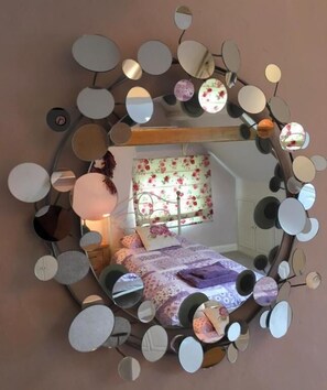 A circular mirror with multiple smaller mirrors attached to it, reflecting a bedroom with a bed, pillows, and a window with floral curtains. - Ceilidh Cottage (York)