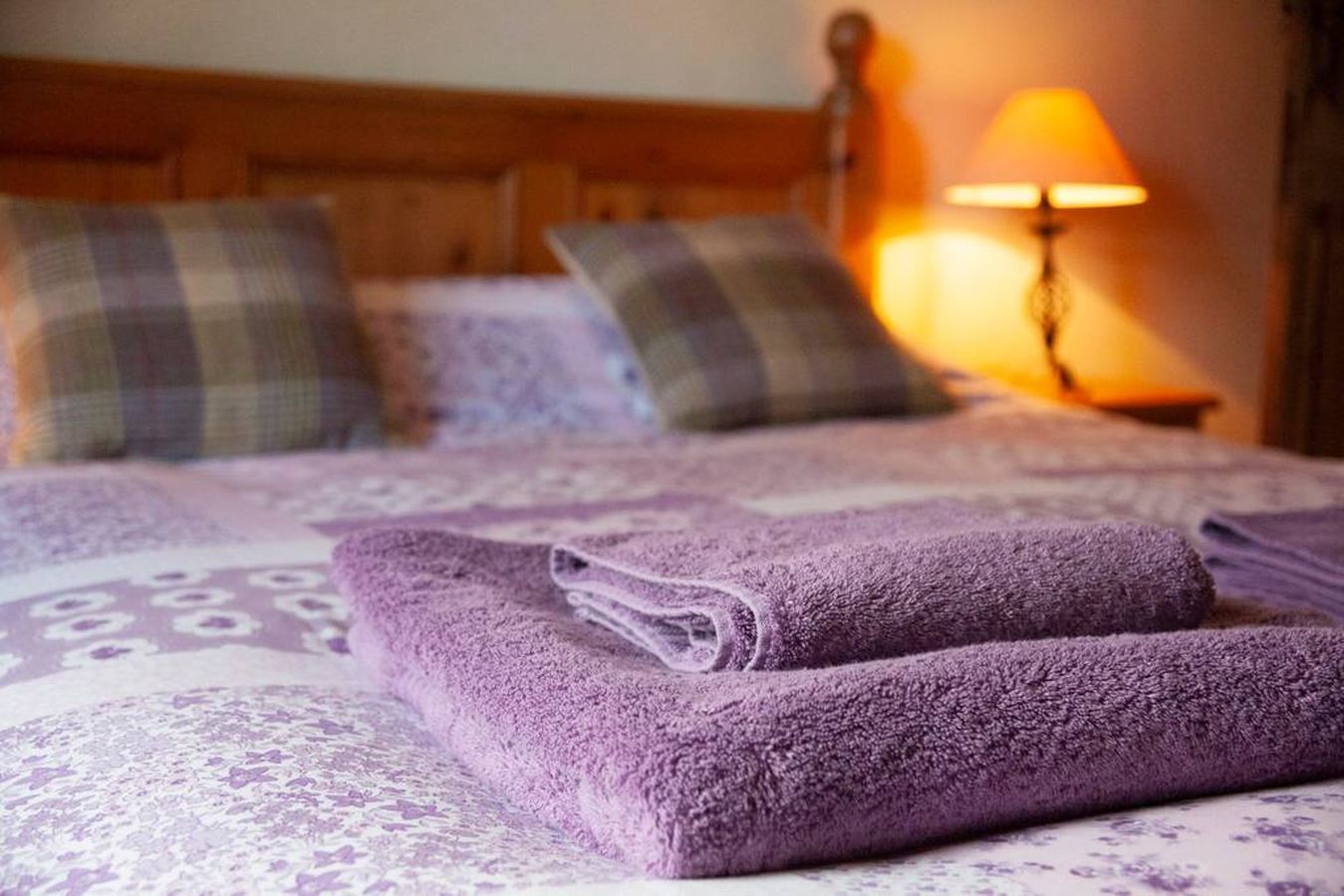 A neatly made bed with purple towels, plaid pillows, and a lamp in the background.
