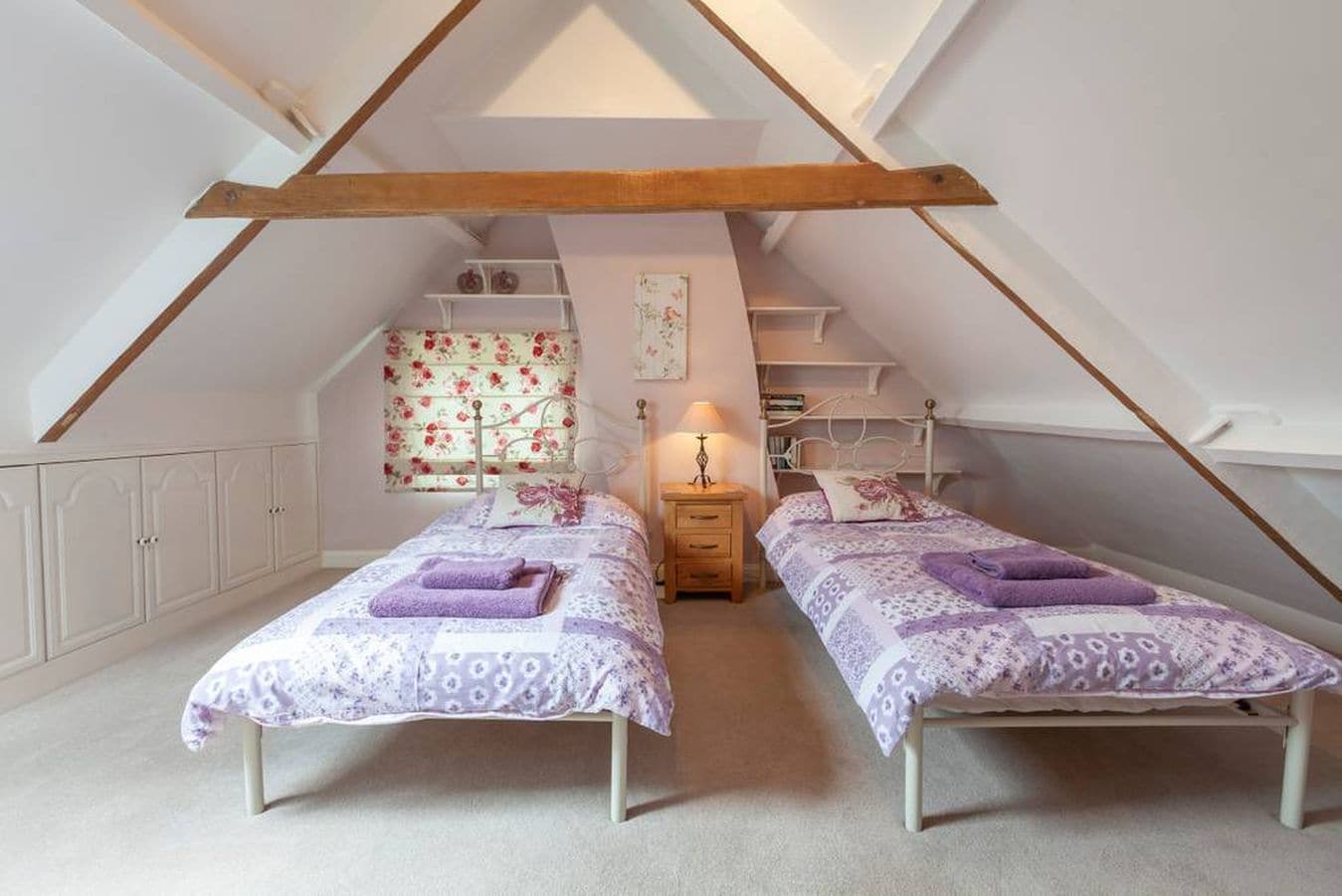 A cozy attic bedroom with two single beds, a small bedside table with a lamp, and a built-in bookshelf.