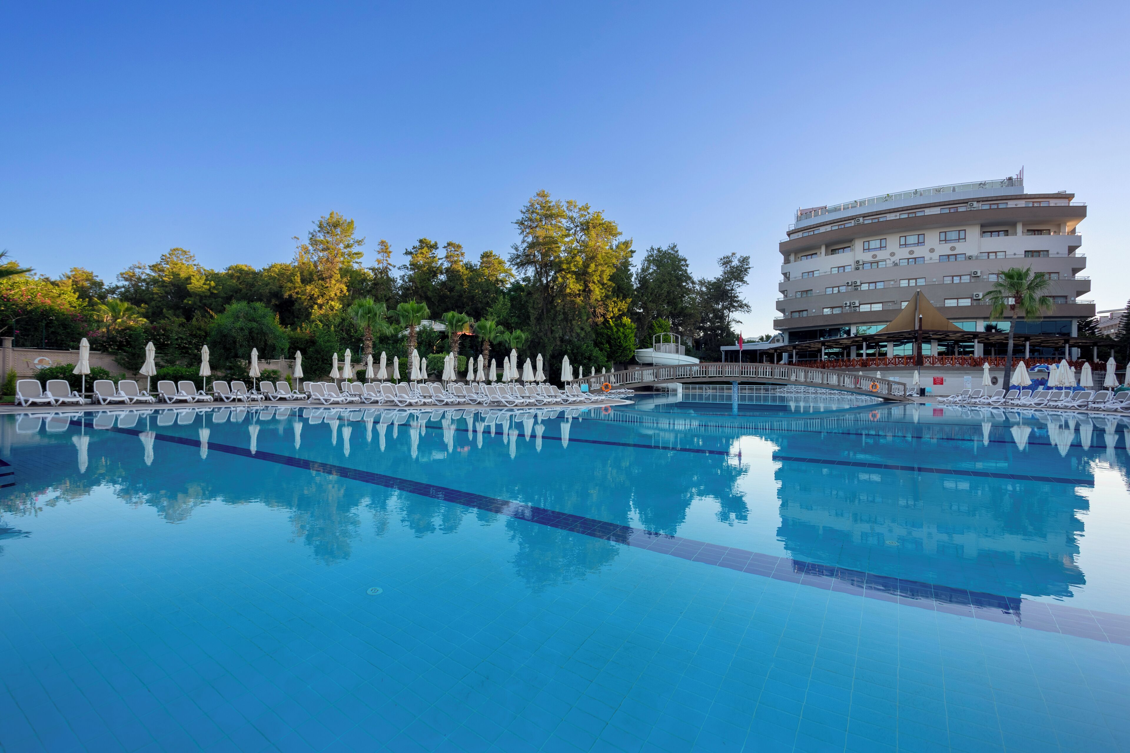 Piscina con sillas de descanso y sombrillas, rodeada de árboles y un edificio de hotel.