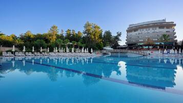 Piscina con sillas de descanso y sombrillas, rodeada de árboles y un edificio de hotel.