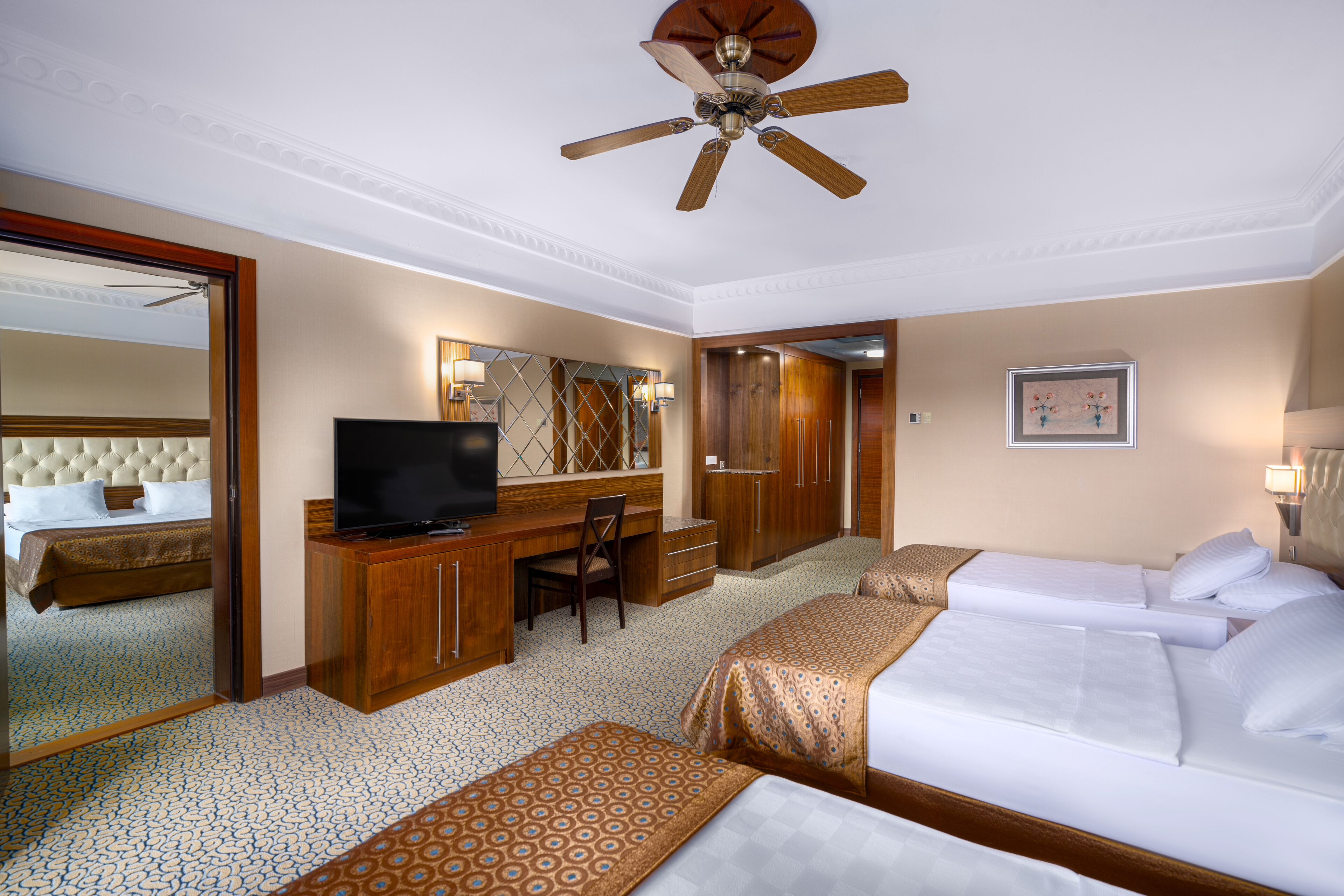 A hotel room with a bed, a desk with a television, a chair, a wardrobe, and a ceiling fan.