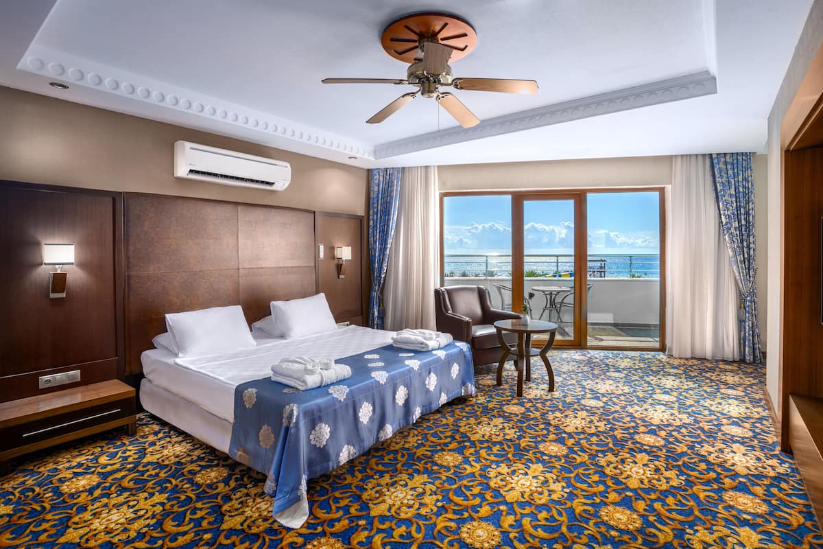 A hotel room with a large bed, a ceiling fan, an air conditioning unit, a balcony with a view of the ocean, and a patterned carpet.