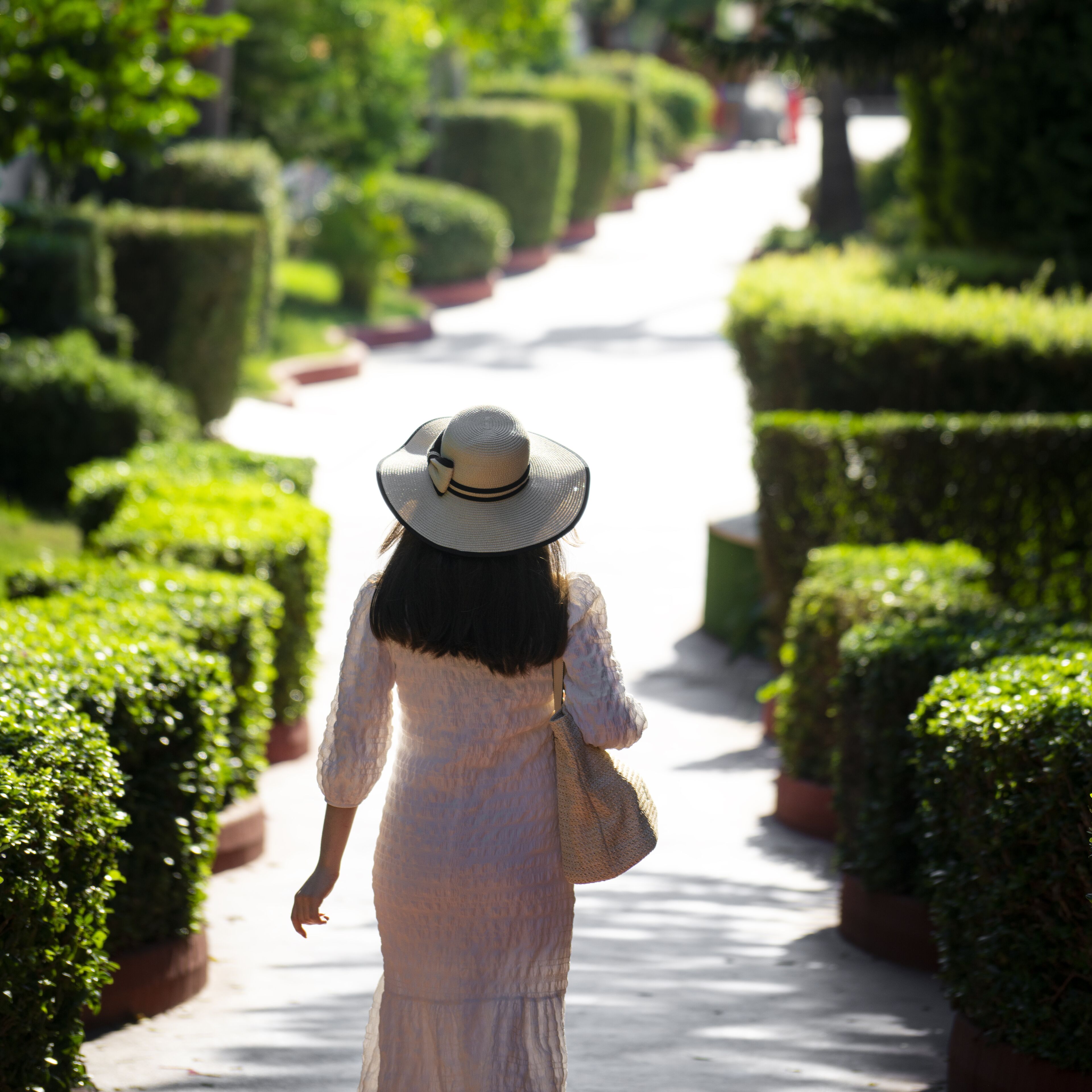 A woman in a white dress and hat walking down a pathway surrounded by neatly trimmed hedges.