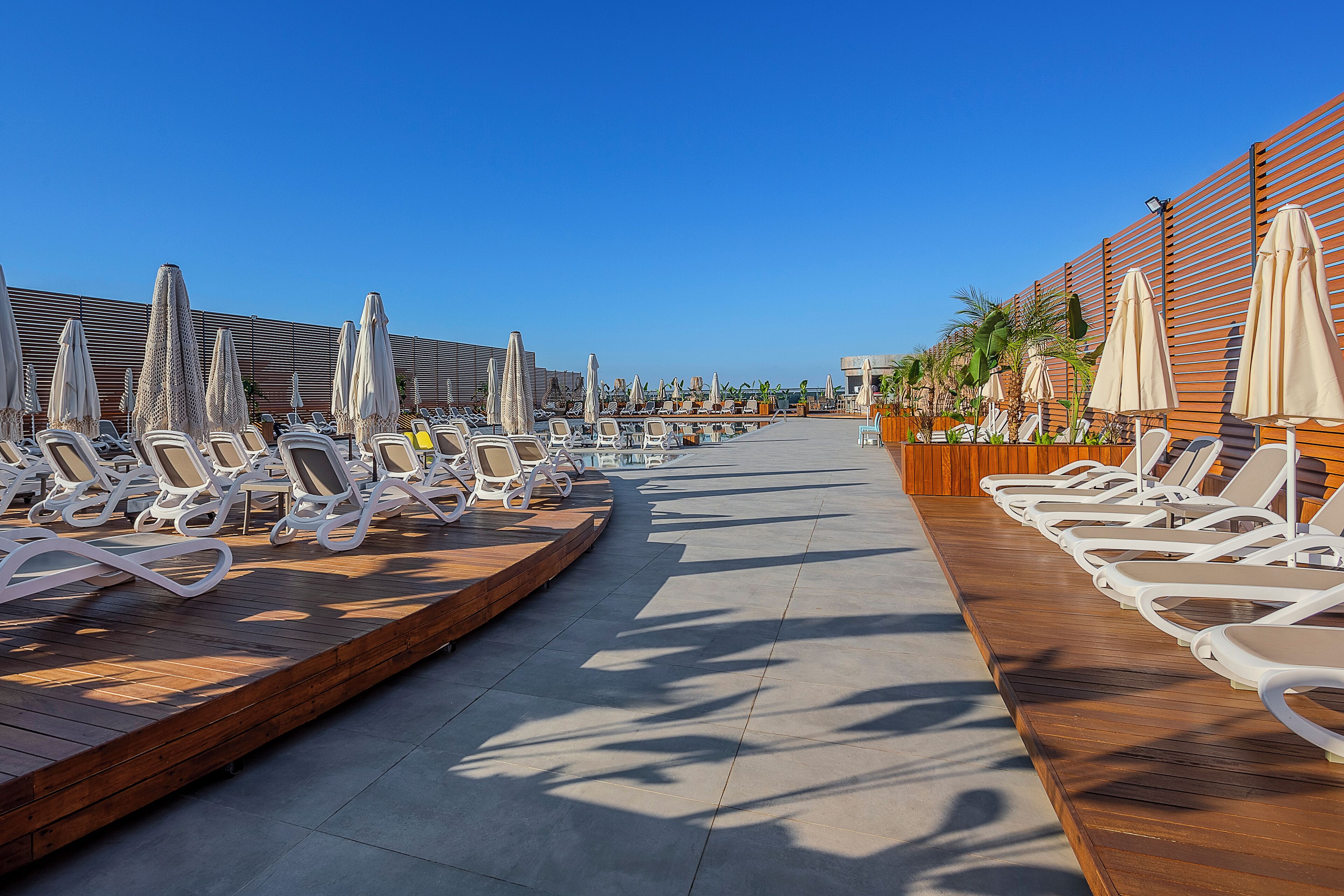 A sunlit outdoor lounge area with rows of white lounge chairs and beige umbrellas, wooden deck, and a clear blue sky.