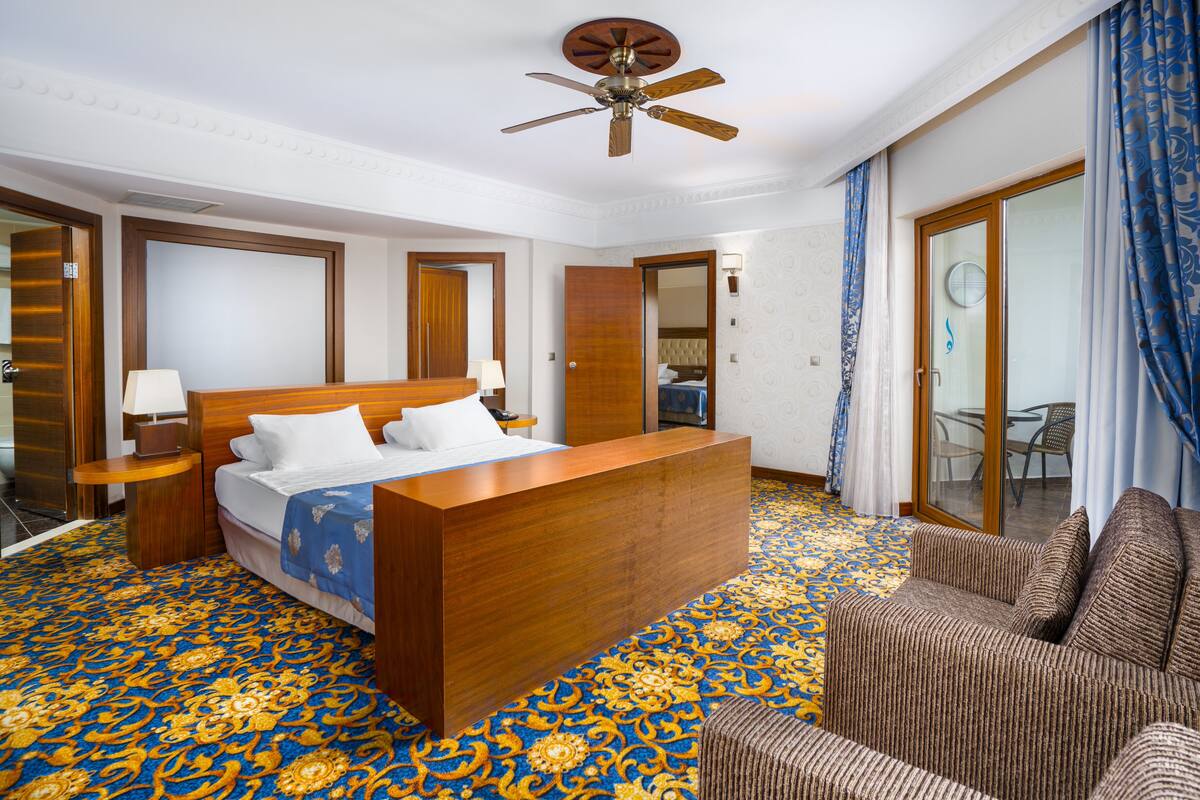 A hotel room with a large bed, a wooden headboard, a ceiling fan, a patterned carpet, and a seating area with a sofa and armchairs.