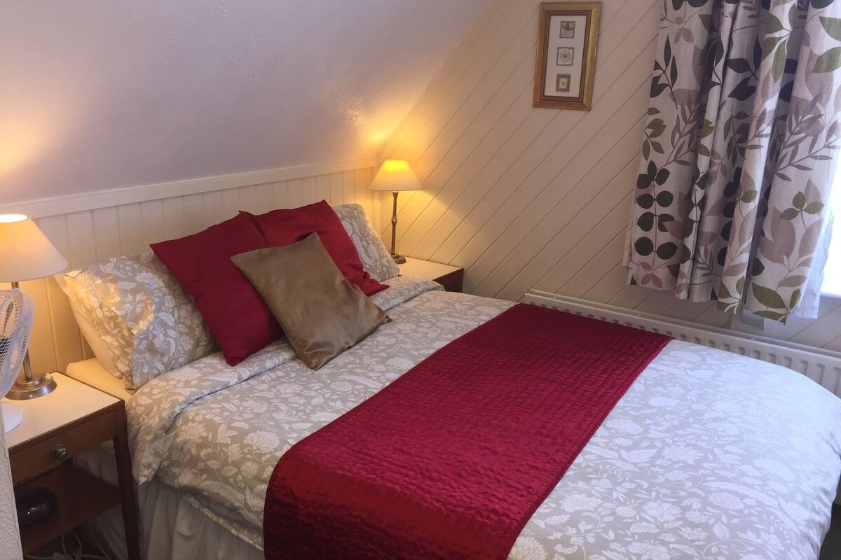 Ein ordentlich bezogenes Bett mit roter Decke, zwei Nachttischlampen und einem blumigem Kissen.