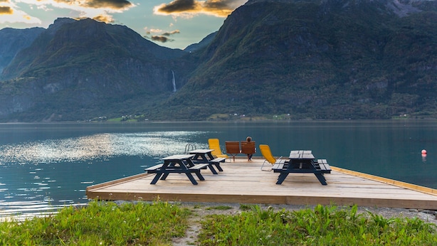 A wooden dock with picnic tables and chairs overlooking a calm lake with mountains in the background.