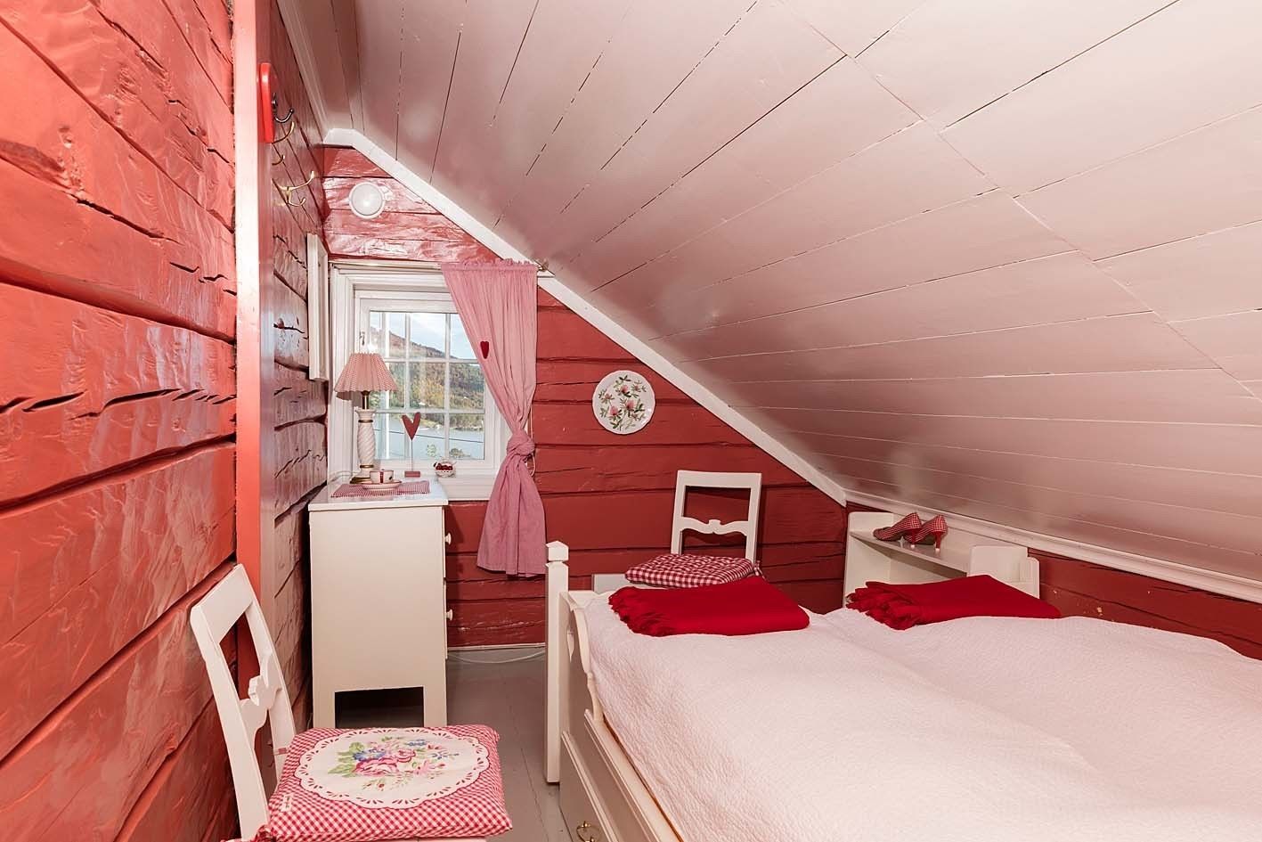 A cozy attic bedroom with a sloped ceiling, a desk with a lamp, a chair, and a bed with red bedding.