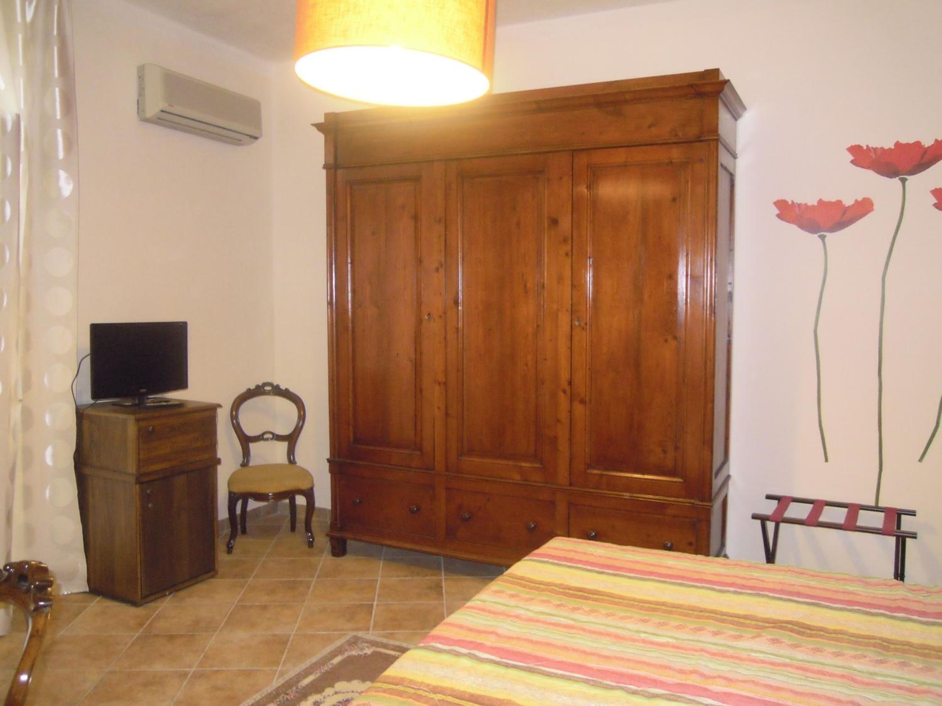 Une chambre avec une armoire en bois, un téléviseur posé sur une petite commode, une chaise et un lit recouvert d’une couverture à rayures.