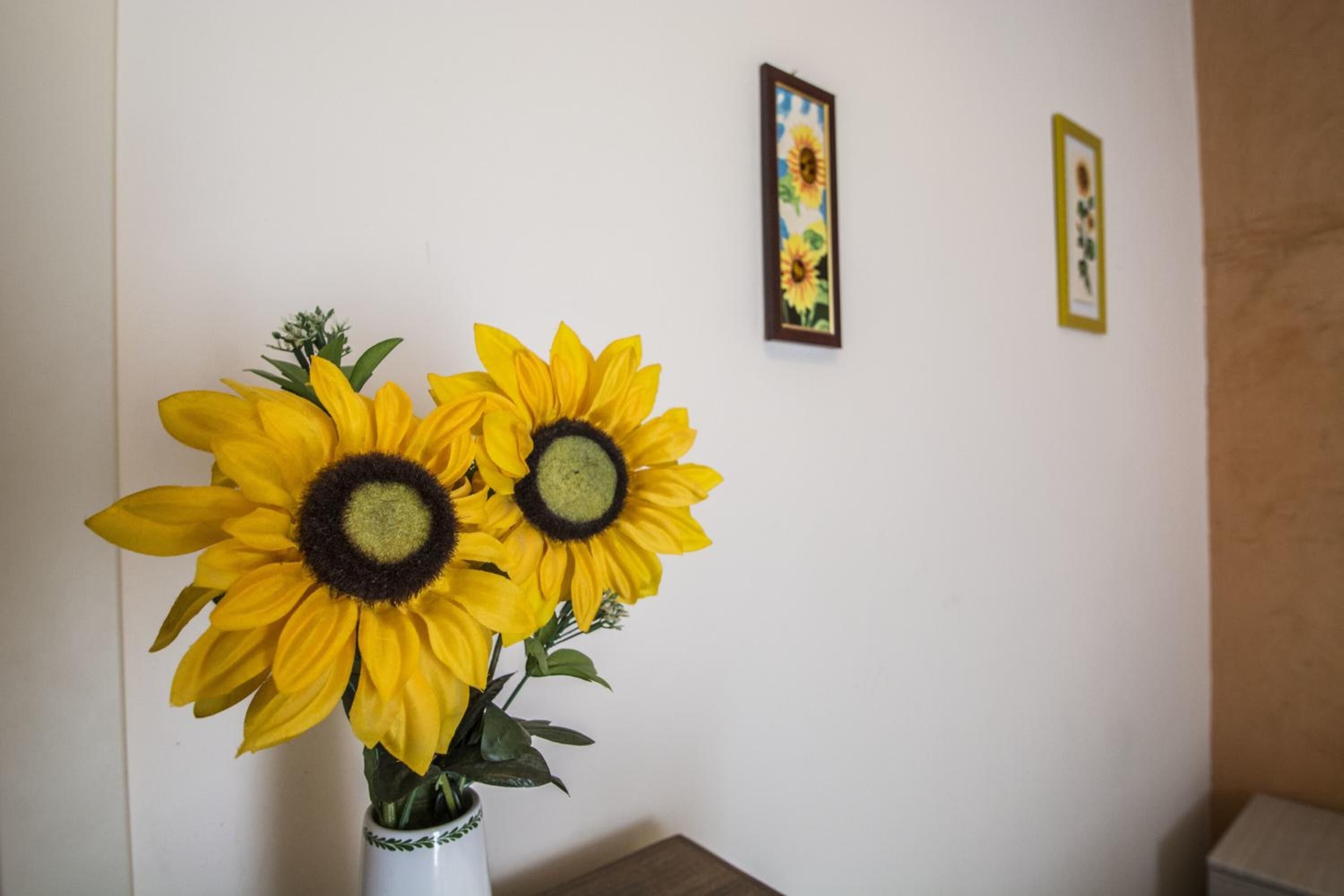 Eine Vase mit zwei Sonnenblumen auf einem Tisch vor einer weißen Wand mit zwei gerahmten Bildern.