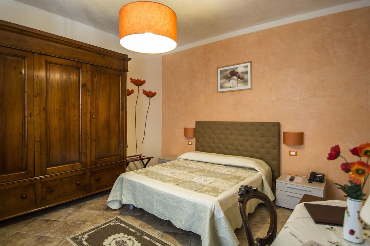 A bedroom with a large wooden wardrobe, a bed with a quilt, a bedside table with a lamp, and a vase with flowers.