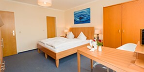 Habitación de hotel con cama, escritorio, silla, mesita de noche, lámpara, un cuadro en la pared y una puerta.
