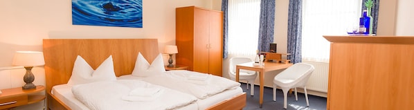 Habitación de hotel con cama, escritorio, silla, armario, lámpara y un cuadro en la pared.