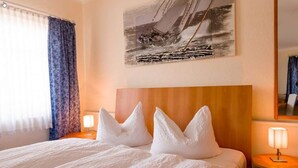 Una habitación de hotel con una cama grande, dos lámparas de noche, un cabecero, una ventana con cortinas y un cuadro de un velero en la pared.