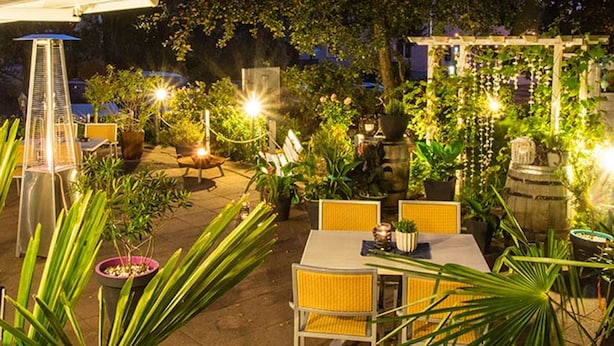 Un área de comedor al aire libre con mesas y sillas, rodeada de plantas en macetas e iluminada con guirnaldas de luces.