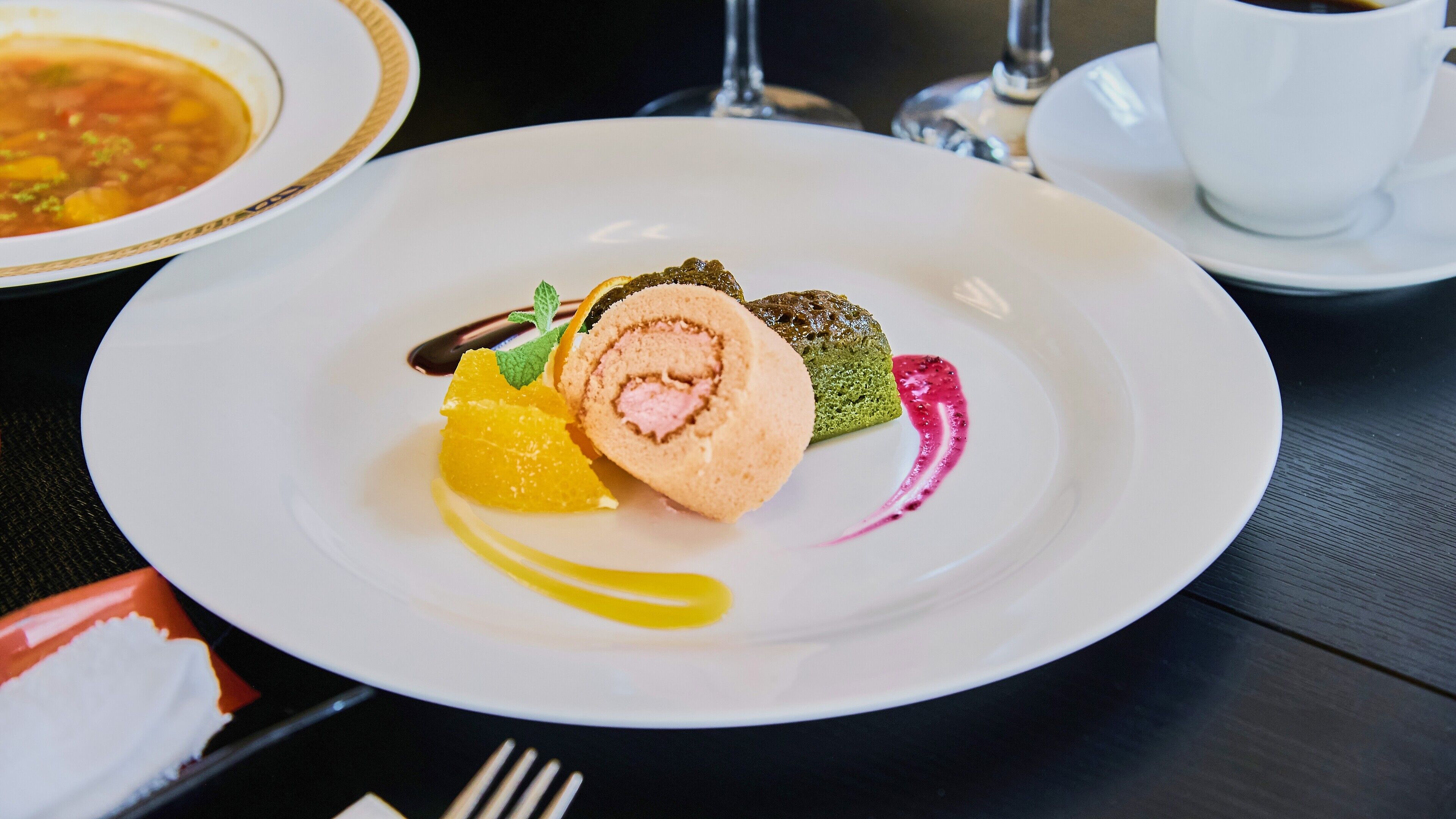 Un plato blanco con un postre que incluye un pastel enrollado, una rodaja de naranja y un elemento verde, acompañado de un poco de salsa.
