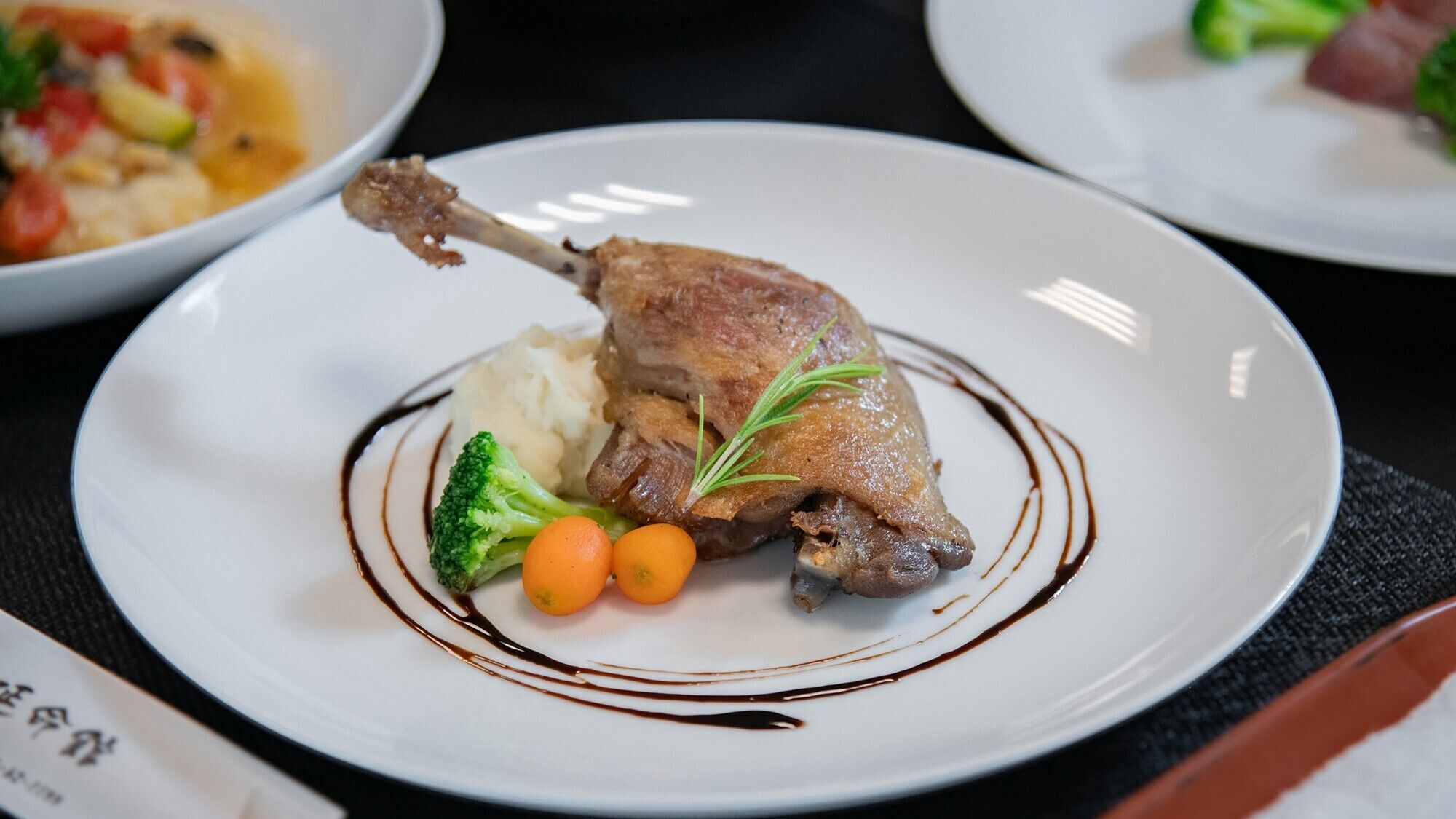 A plate with a roasted duck leg, mashed potatoes, broccoli, and carrots, garnished with a sprig of rosemary and a chocolate drizzle.