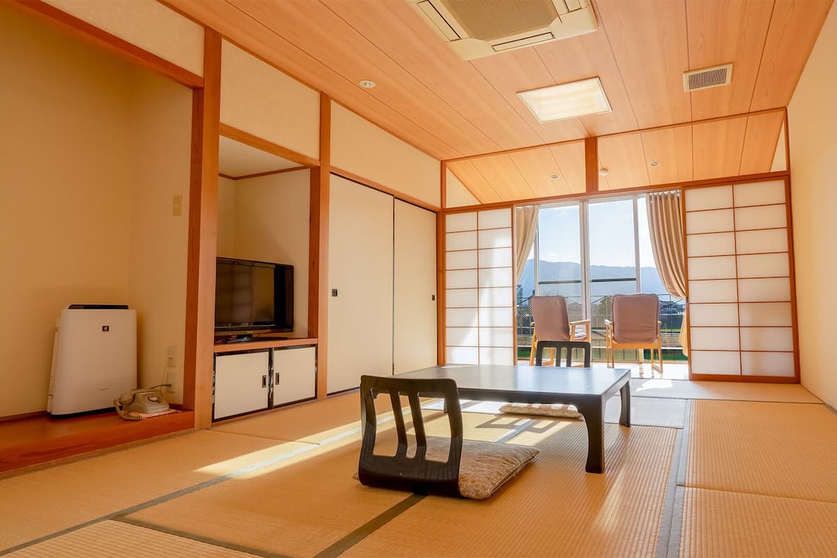 A traditional Japanese room with tatami flooring, a television, a refrigerator, and a sliding door leading to a balcony with a view of the mountains.