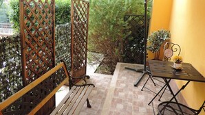 Un espace patio aménagé avec un banc en bois, une table en métal entourée de chaises et un parasol blanc offrant de l’ombre.