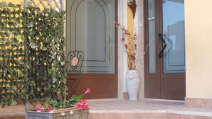 Une entrée d’immeuble ornée d’un treillage en bois, d’un grand pot de fleurs contenant une plante aux fleurs roses et d’un vase décoratif avec des branches séchées.