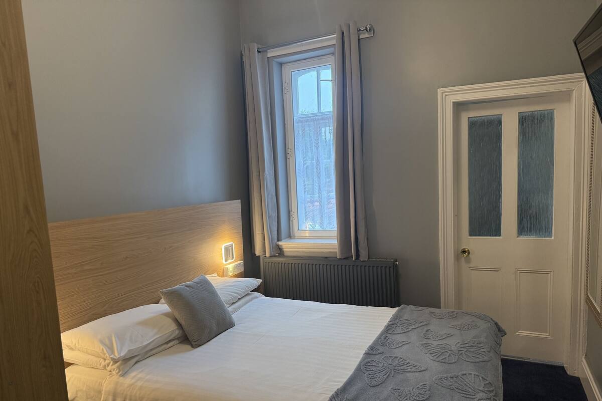 A hotel room with a bed, a window with curtains, a radiator, and a door.