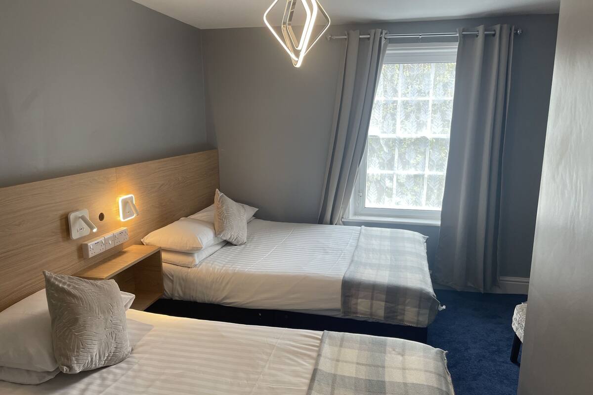 A hotel room with two beds, a wooden headboard, bedside lamps, a window with curtains, and a blue carpet.